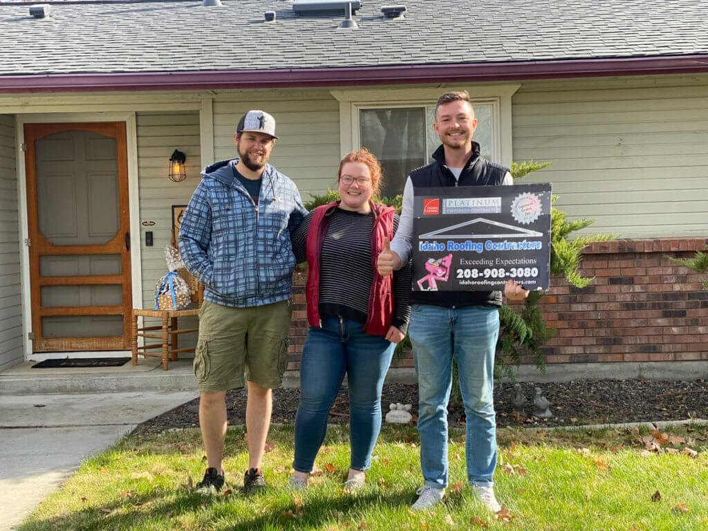 Three people standing in front of a house, holding a sign for Idaho Roofing Contractors.