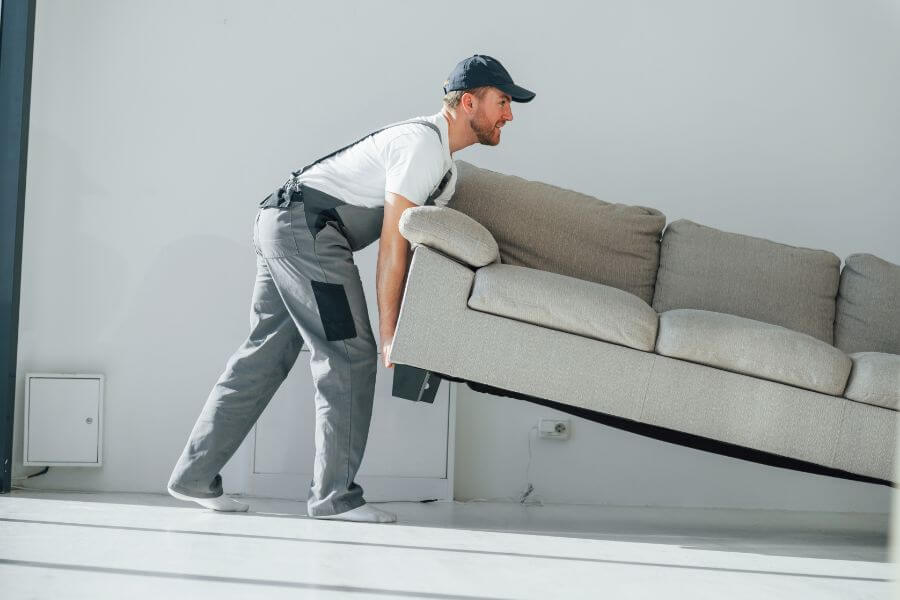 A mover in uniform lifts a beige sofa inside a well-lit room.