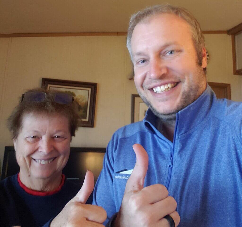 Two smiling people, an older woman and a man in a blue jacket, giving thumbs up while standing together indoors.