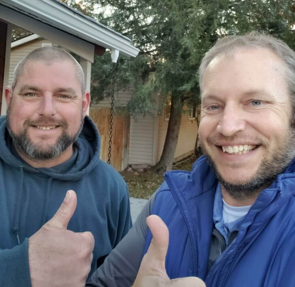 Two men giving thumbs up and smiling, standing outdoors with trees and houses in the background.
