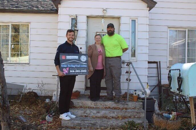 Three people standing on steps in front of a house, holding a sign for Idaho Roofing Contractors.