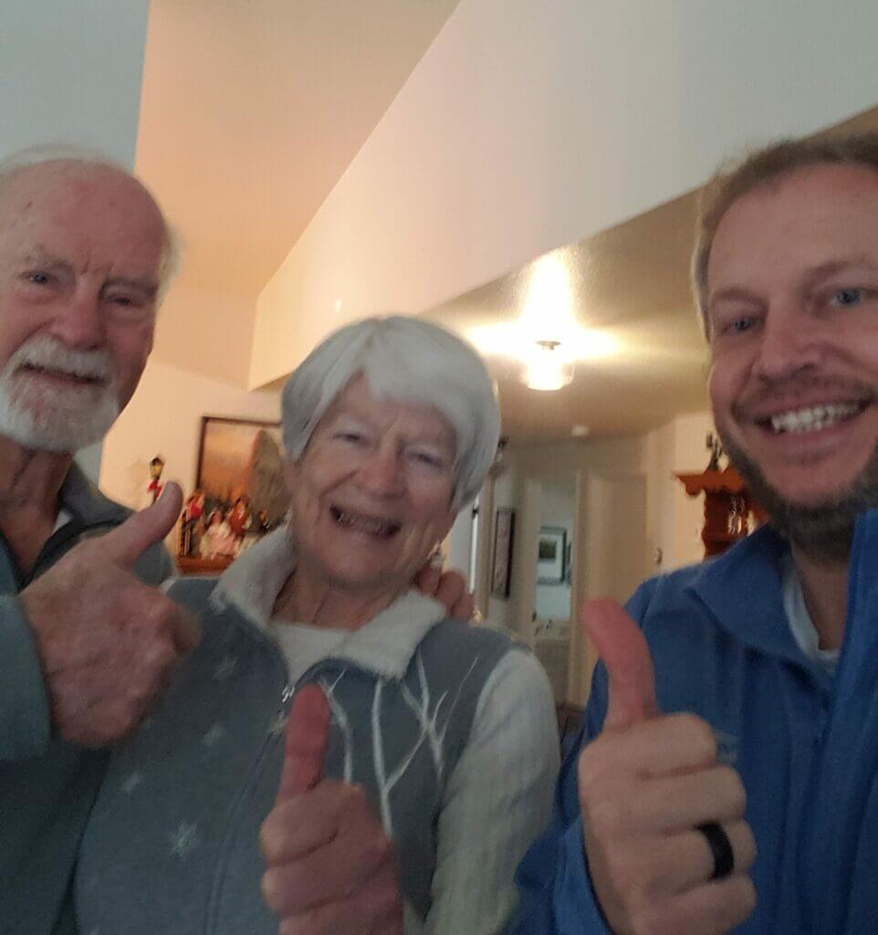 Two smiling elderly people and a man in a blue jacket giving thumbs up while standing together indoors.