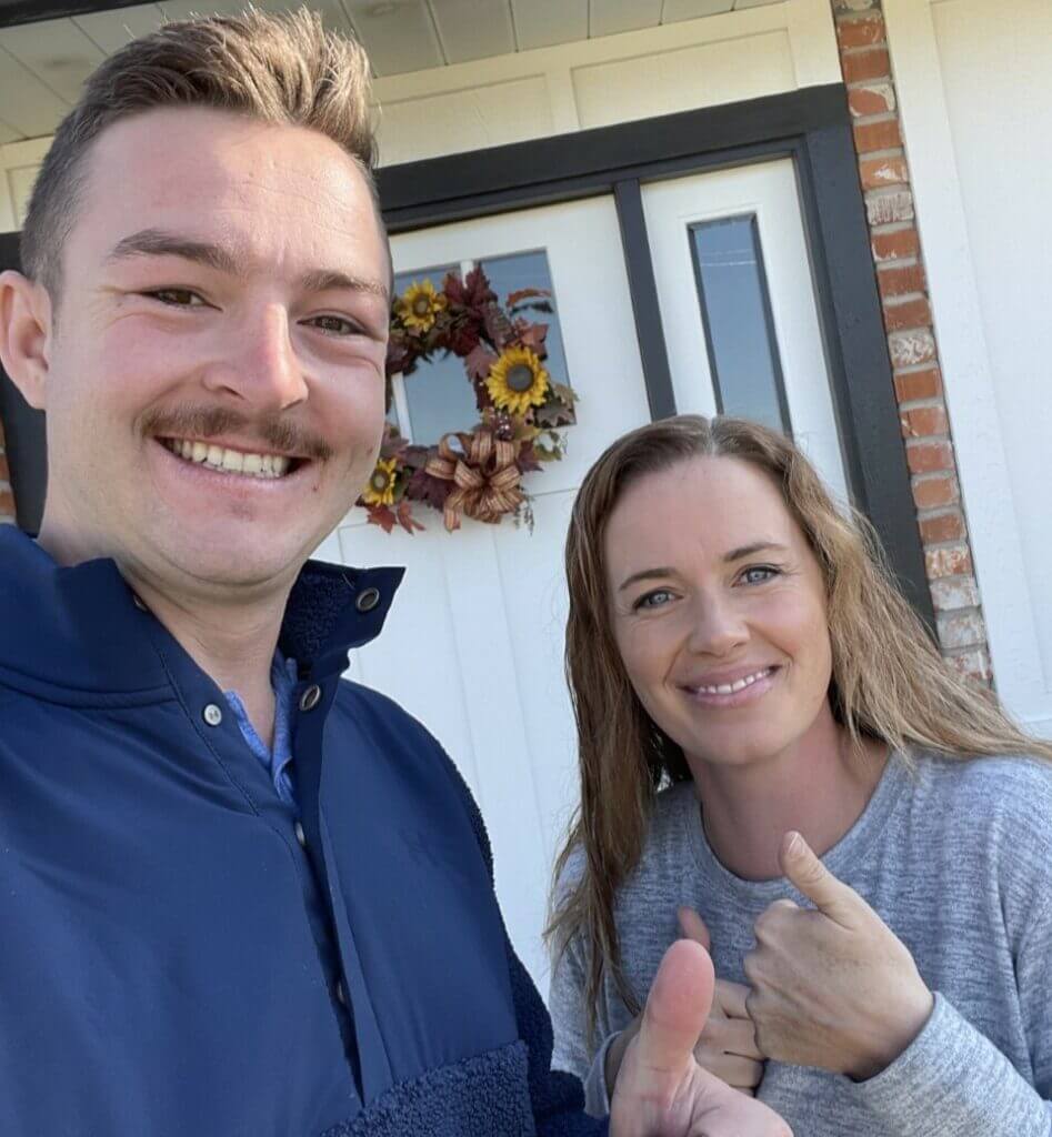 Man and woman standing outside a home, both smiling and giving thumbs up, with a decorative wreath on the door behind them.