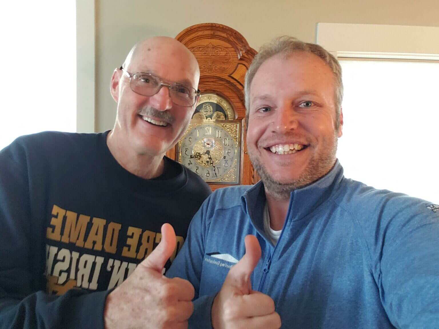Two smiling men giving thumbs up in front of a wooden clock.