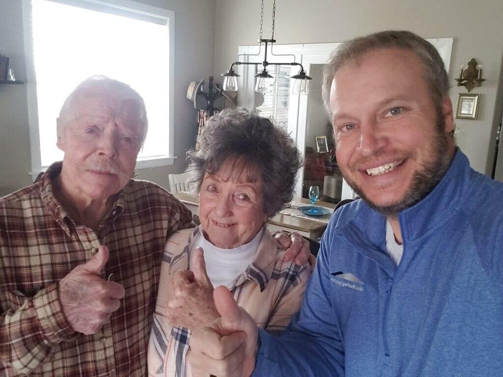 Two elderly people and a man in a blue jacket giving thumbs up while standing together indoors.