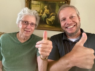 Elderly woman and man in a black shirt giving thumbs up while sitting together indoors.