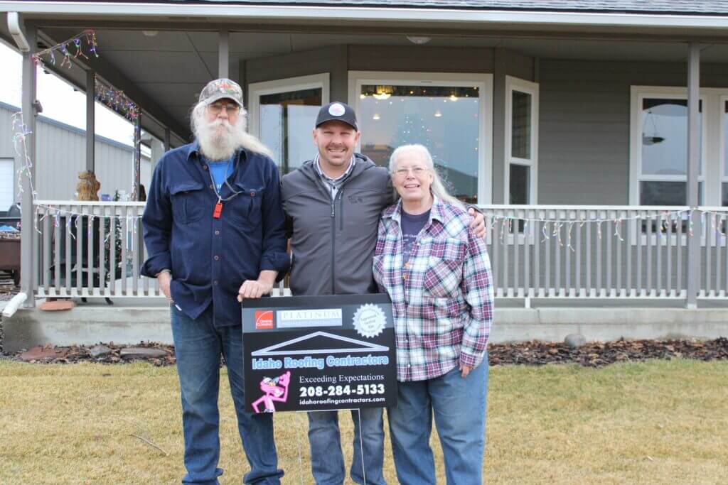 Two men and a woman standing in front of a house, holding a sign for Idaho Roofing Contractors.