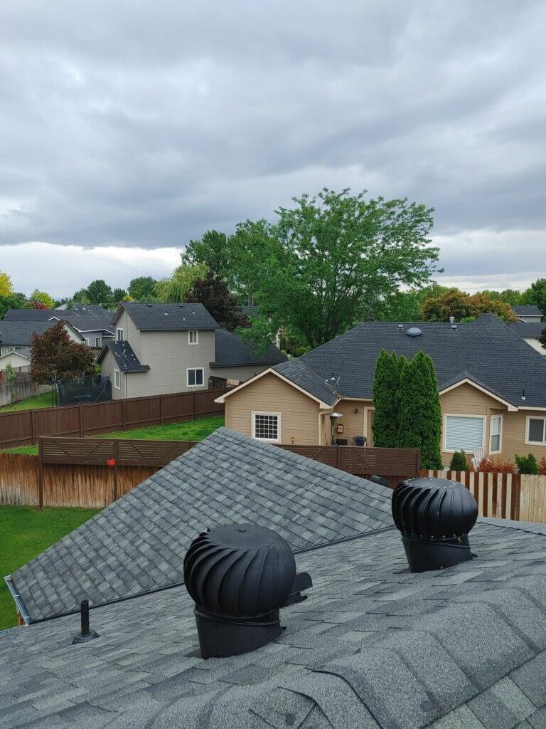 A roof with newly installed shingles featuring two black turbine vents against a background of neighborhood houses and trees.