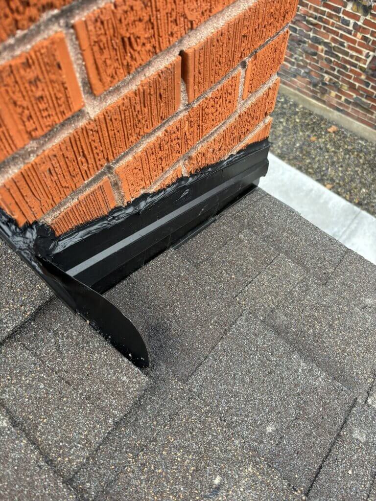 Close-up of black step flashing installed where roof meets brick chimney, sealing against water intrusion.