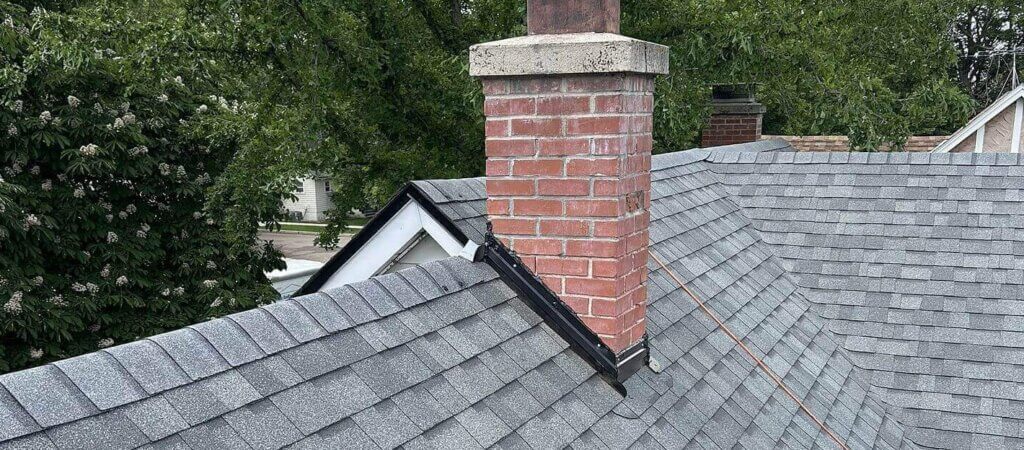 A close-up of a brick chimney sealed with step flashing, intersecting slopes, and a gray shingle roof with a steep pitch.