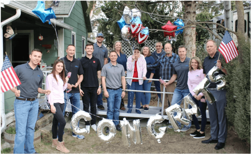 Team and community members celebrate outside a home with a military family, holding flags and “Congrats” balloons after completing a roof replacement in Idaho.