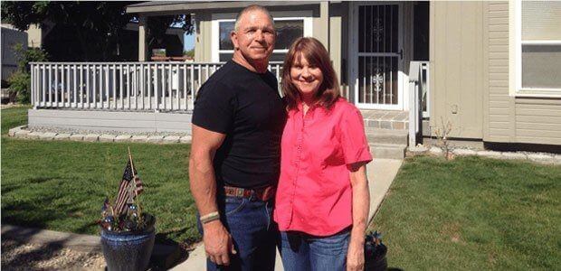 A smiling couple stands in front of their home on a sunny day, with small American flags displayed in planters.