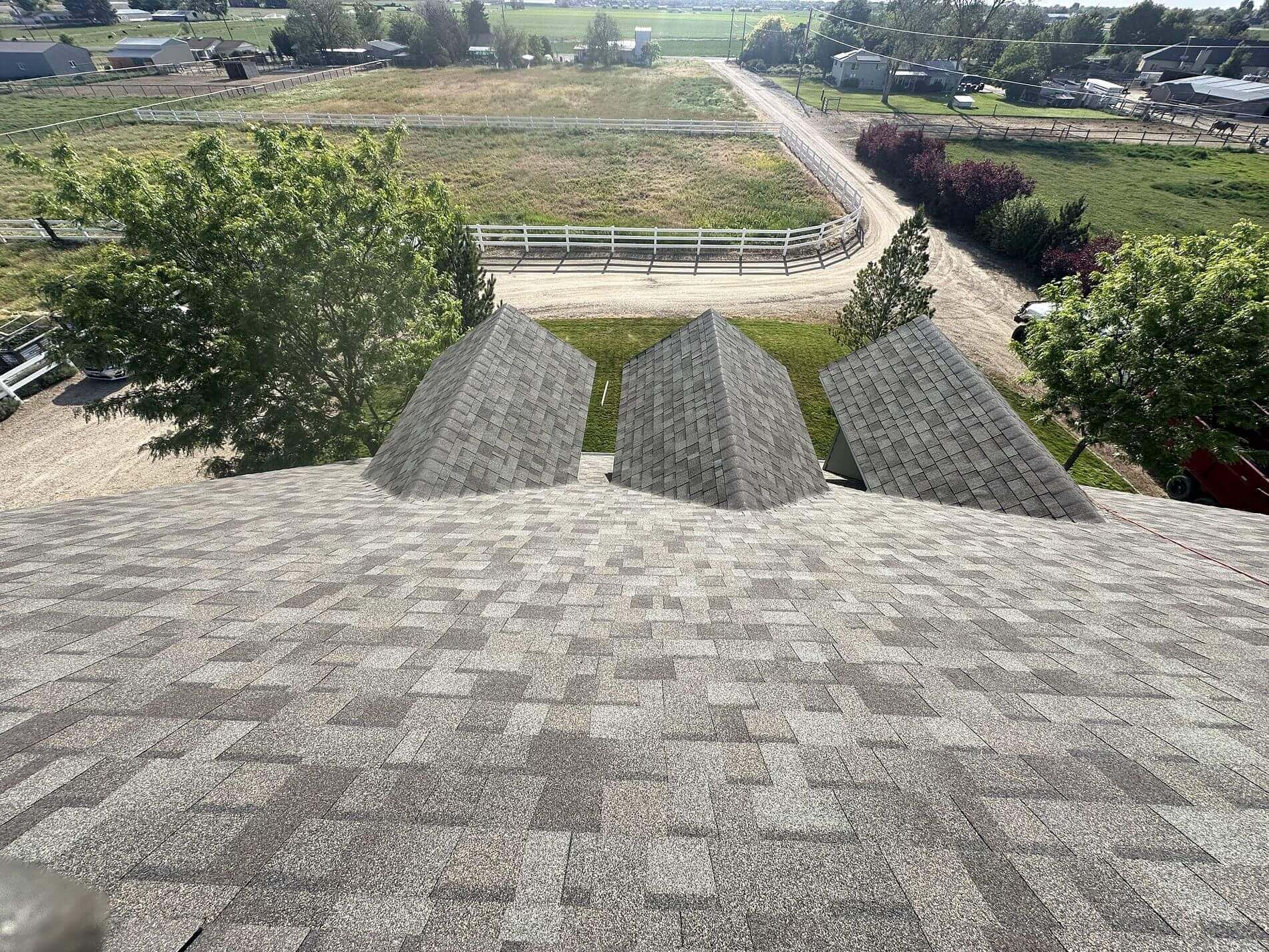 Three gable projections of a shingle roof face a dirt driveway and enclosed agricultural land in a tranquil rural area.