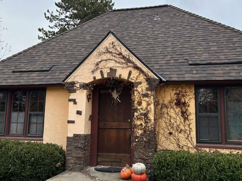 Cottage-style stucco home with dark brown shingle roof, wooden front door, and decorative fall pumpkins.