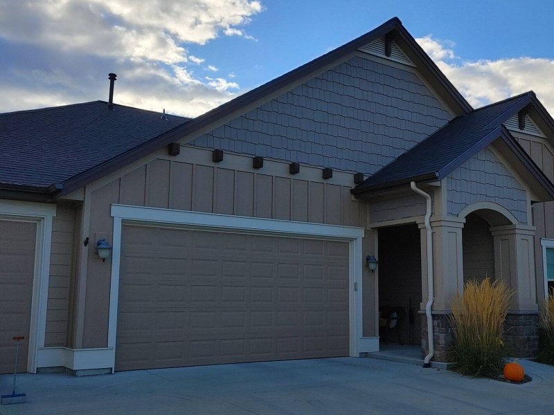 Suburban home with dark roof, tan siding and two-car garage beneath partly cloudy sky.